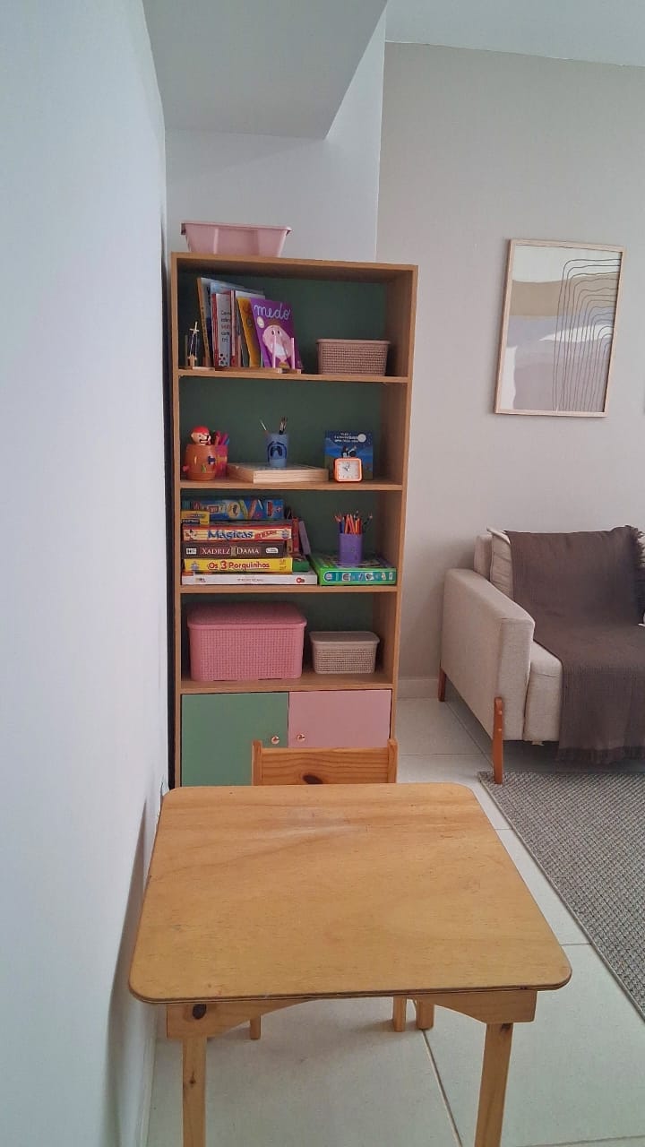 Canto do consultório com uma mesinha de madeira, cadeira infantil e uma estante com livros, jogos e caixas organizadoras em tons de verde e rosa.