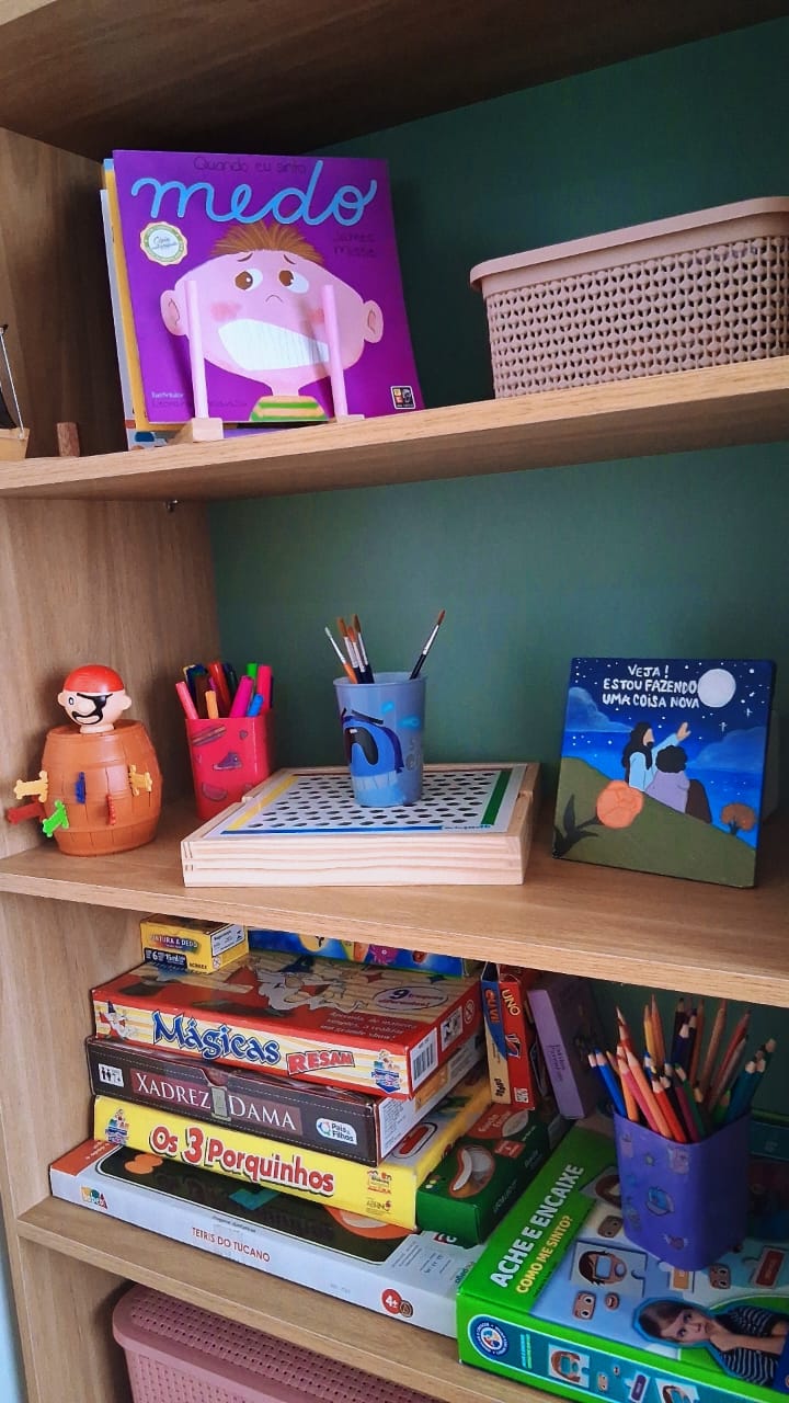 Prateleiras com livros infantis sobre emoções, jogos de tabuleiro, lápis de cor, pincéis e brinquedos.