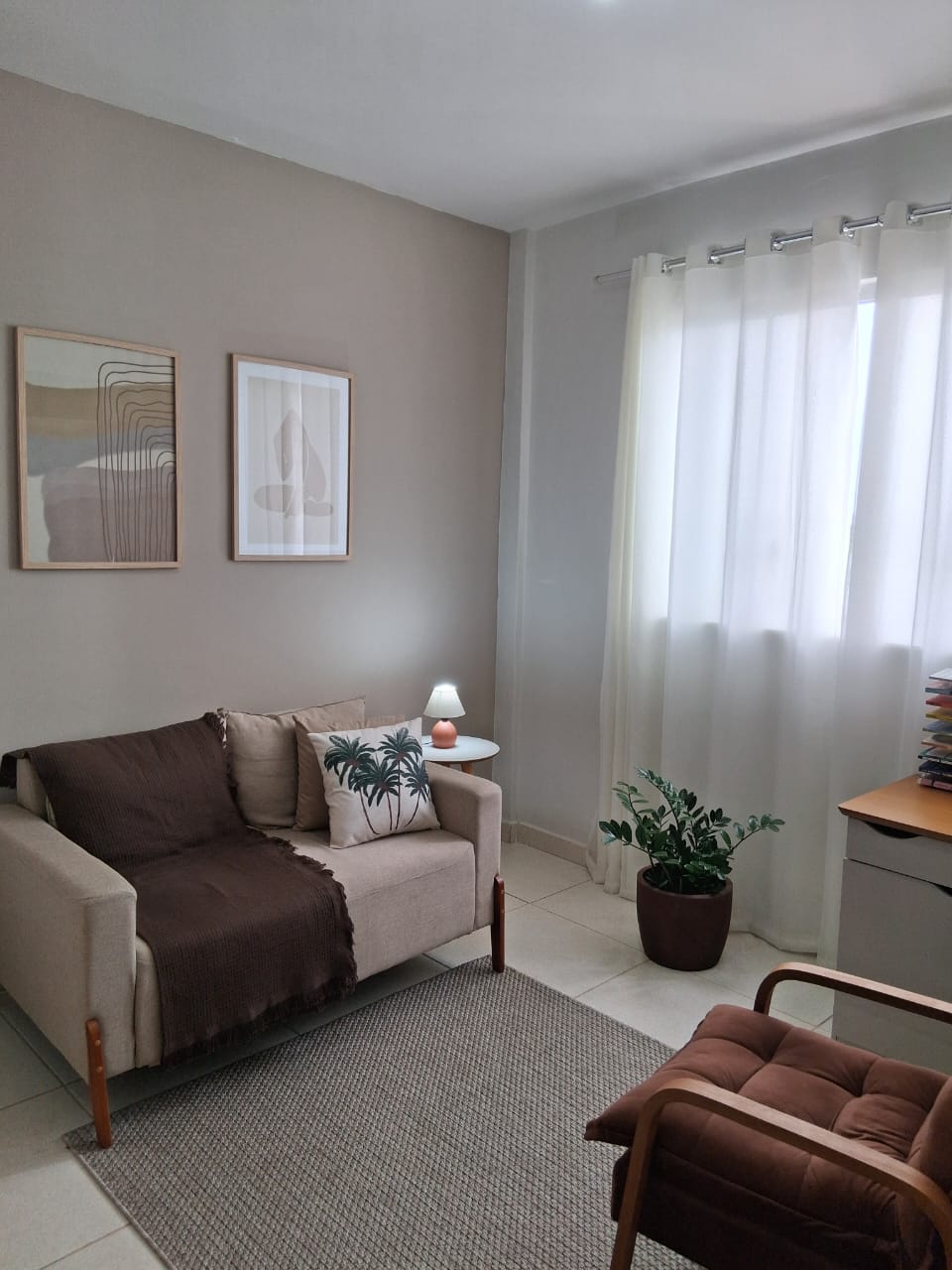 Sala de atendimento com sofá claro, mantas, almofadas, quadros de tons neutros, planta e cortina translúcida.