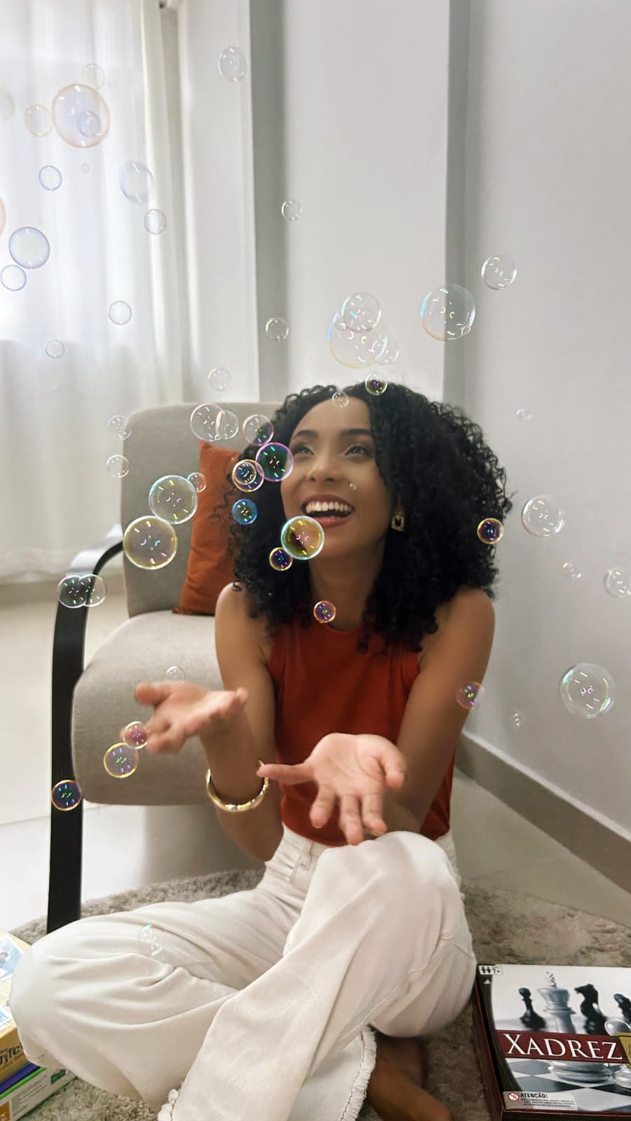 Thalita sentada no chão de um ambiente claro, rindo, com as mãos abertas entre bolhas de sabão.