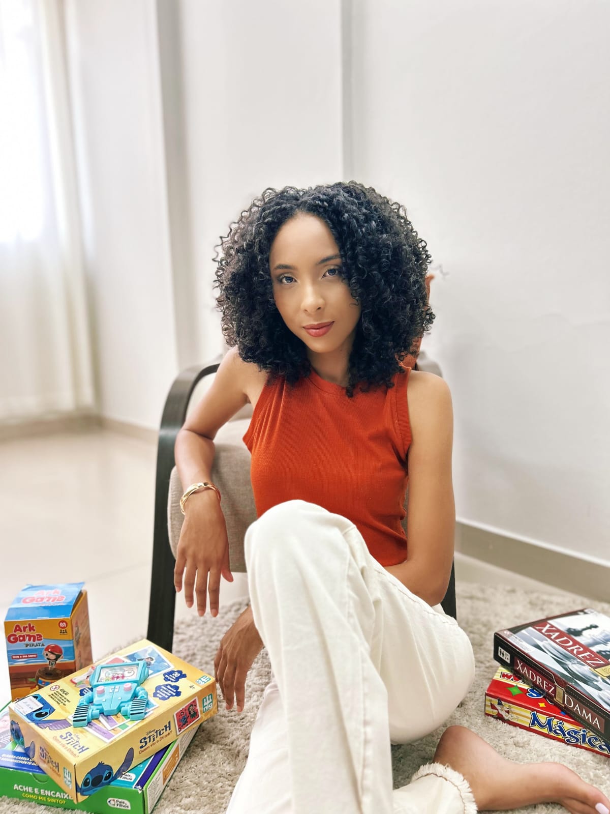 Thalita sentada no tapete, encostada em uma poltrona, com caixas de jogos ao seu redor.