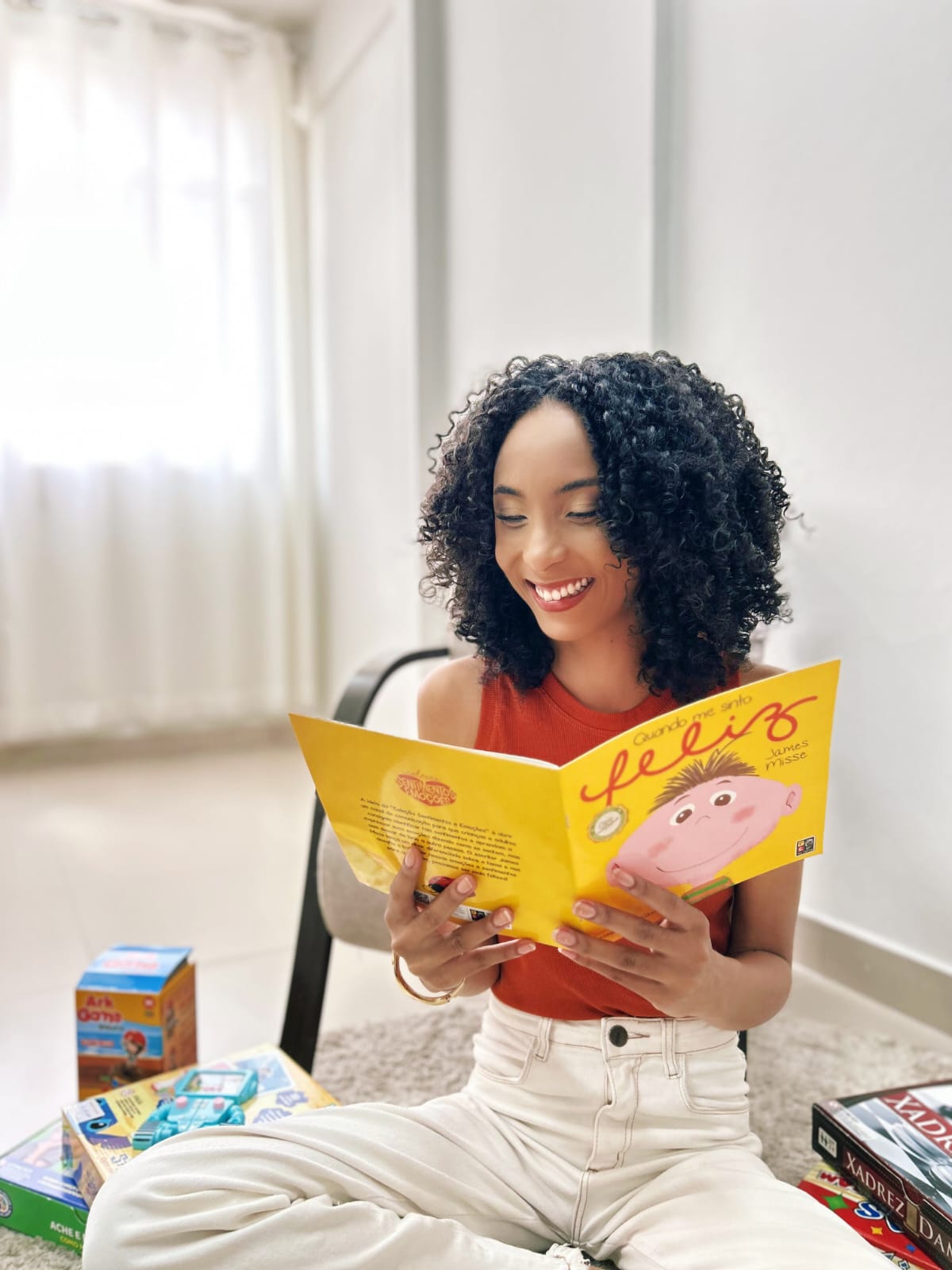 Thalita sorrindo enquanto lê o livro infantil “Quando me sinto feliz”, sentada no tapete com jogos ao lado.