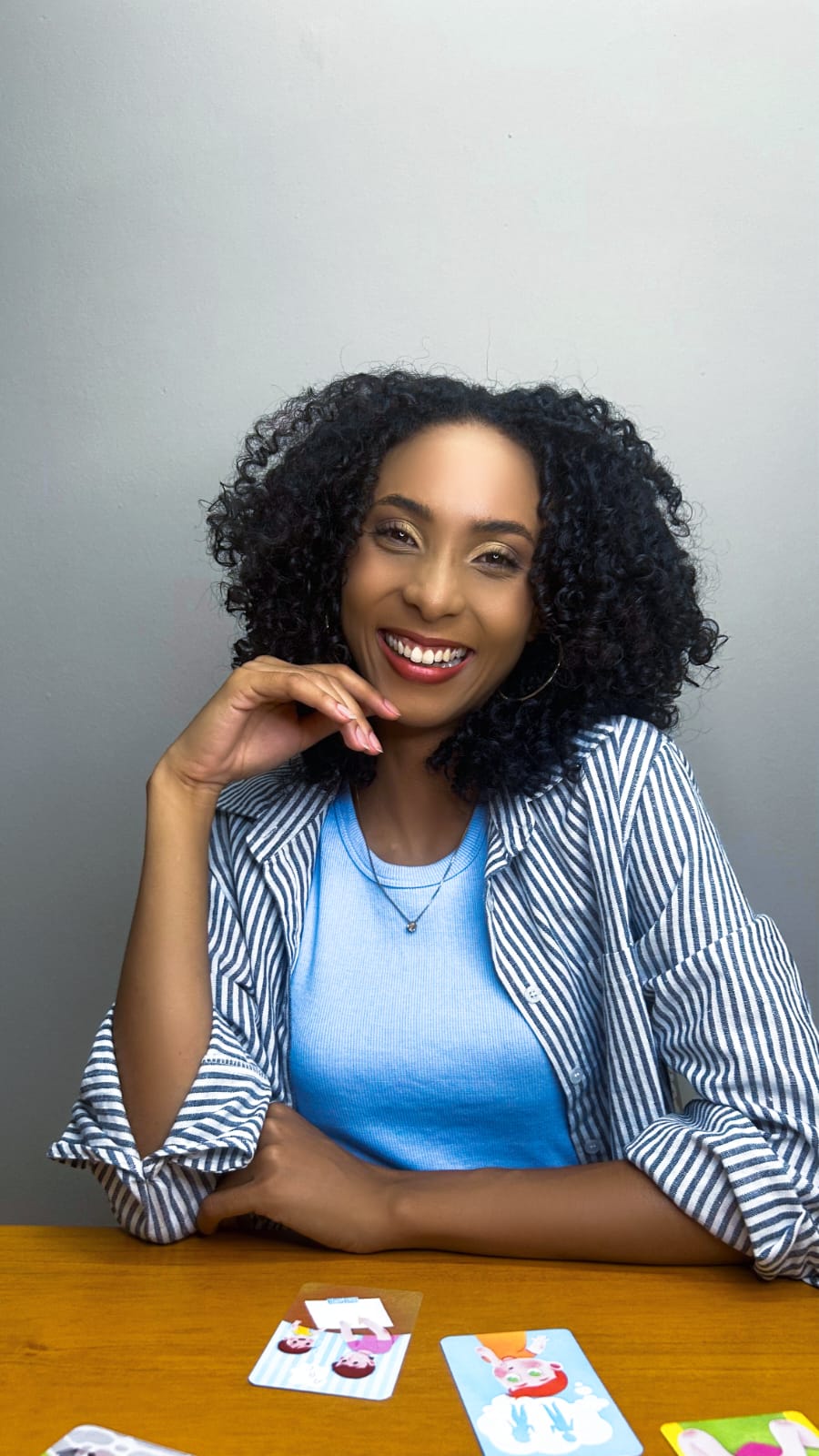 Thalita Gonçalves sorrindo, sentada à mesa com blusa azul e camisa listrada, apoiando o rosto na mão, com cartas ilustradas à sua frente.