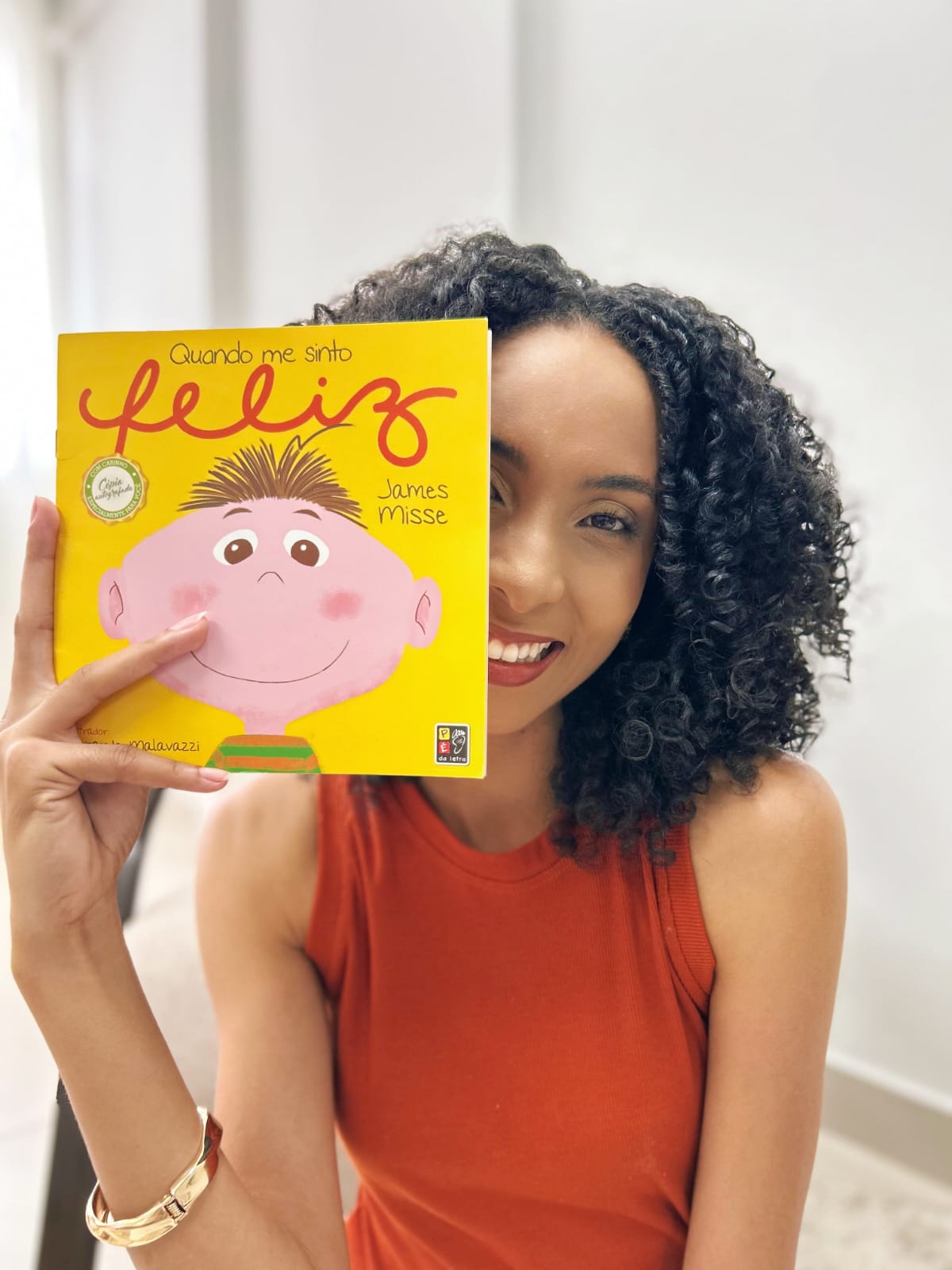 Thalita sorrindo, segurando ao lado do rosto o livro infantil “Quando me sinto feliz”.