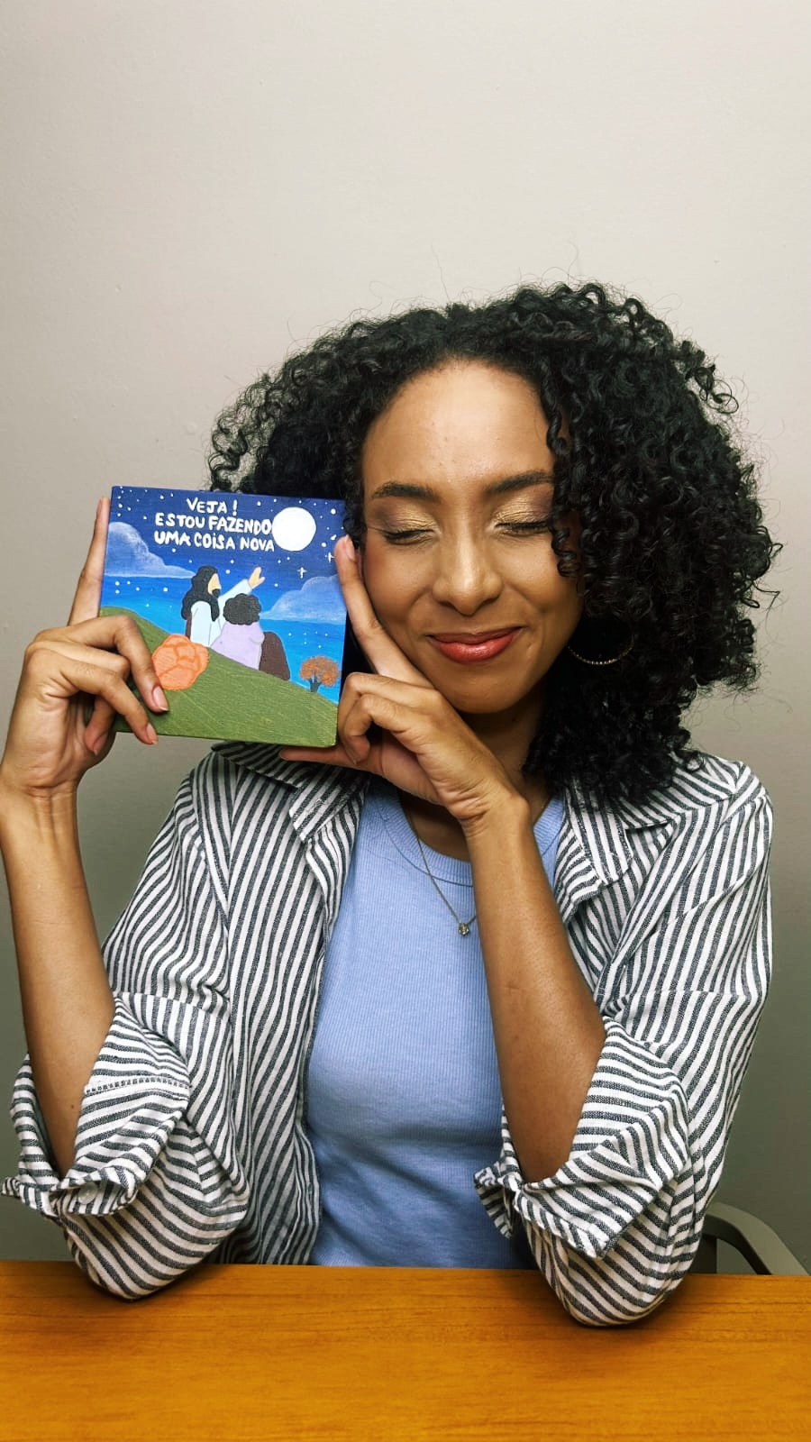 Thalita sorrindo de olhos fechados, segurando ao lado do rosto um livro infantil com a frase “Veja! Estou fazendo uma coisa nova”.
